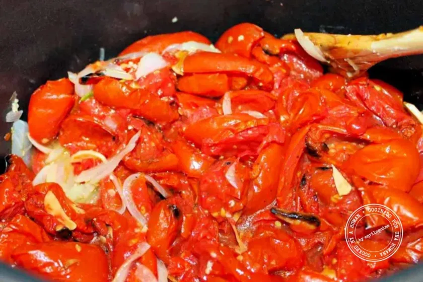 Roasted tomatoes and thinly sliced onions being stirred together in a pot with a wooden spoon.