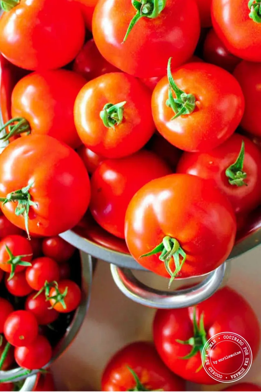 Un colador metálico lleno de tomates de un rojo intenso, con más tomates y tomates cherry a la vista en recipientes cercanos.