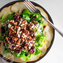 Una ensalada de peras, estilo carpaccio con nueces pecanas, arándanos rojos secos y queso azul,con un tenedor en el plato.