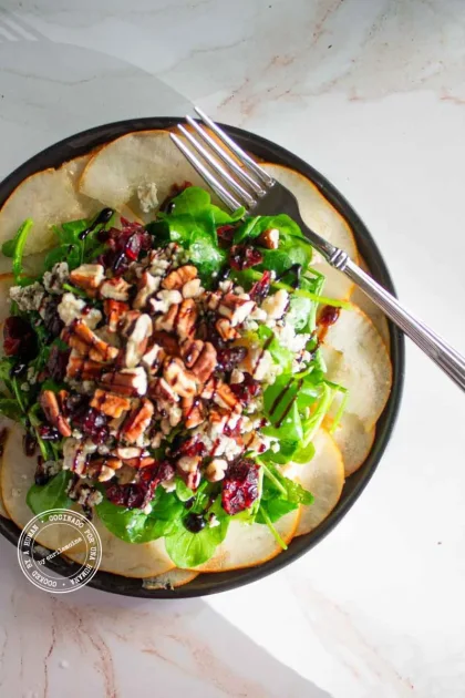 Una ensalada de peras, estilo carpaccio con nueces pecanas, arándanos rojos secos y queso azul,con un tenedor en el plato.
