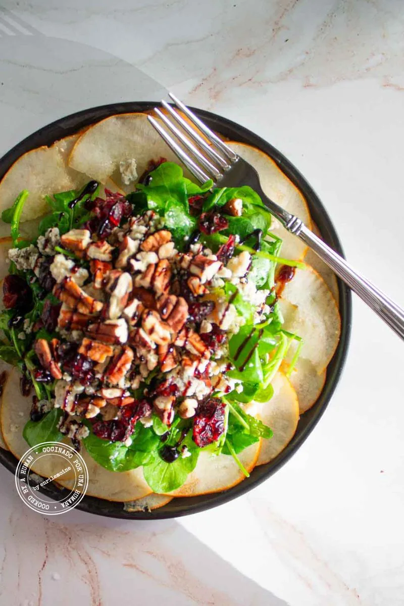 Una ensalada de peras, estilo carpaccio con nueces pecanas, arándanos rojos secos y queso azul,con un tenedor en el plato.