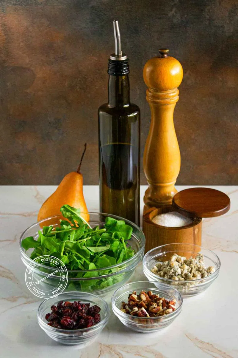 Ingredientes para una ensalada que incluye una pera, rúcula, queso azul, arándanos rojos secos, nueces pecanas, aceite de oliva, sal y pimienta.