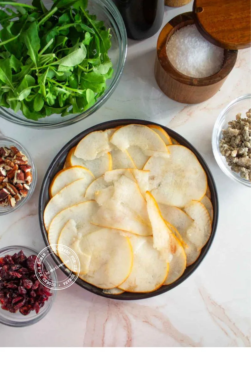 Un plato negro con peras cortadas en rodajas finas, rodeado de cuencos pequeños con rúcula, nueces pecanas, arándanos rojos secos, una mezcla de especias y un salero.