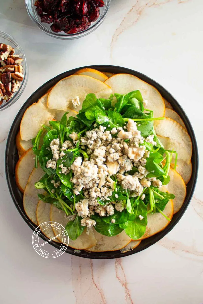 Una ensalada de peras cortadas en rodajas, con rúcula y queso azul en un plato, con cuencos de nueces pecanas y arándanos rojos secos al lado.