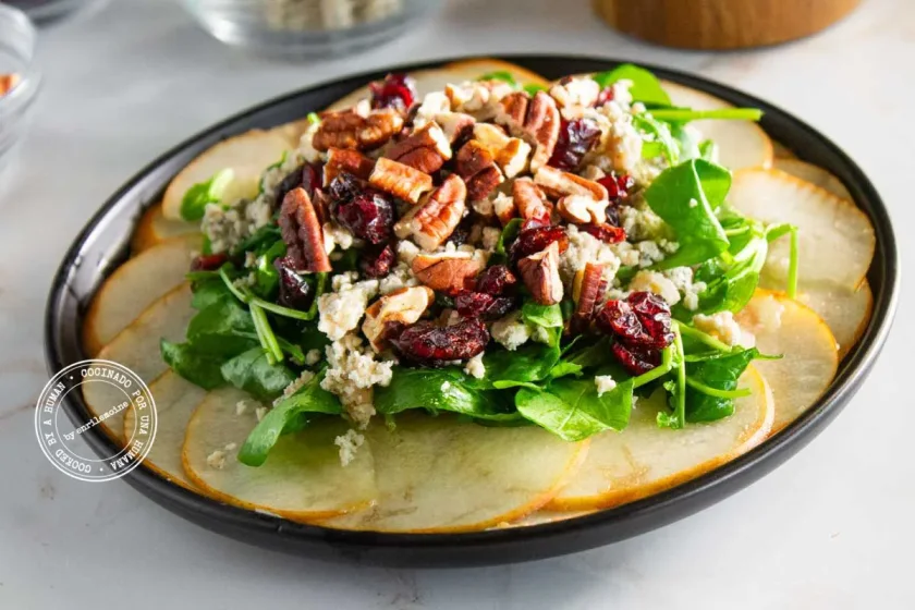 Una ensalada con finas rodajas de pera, verduras de hoja verde, queso azul, nueces pecanas y arándanos rojos secos en un plato negro.