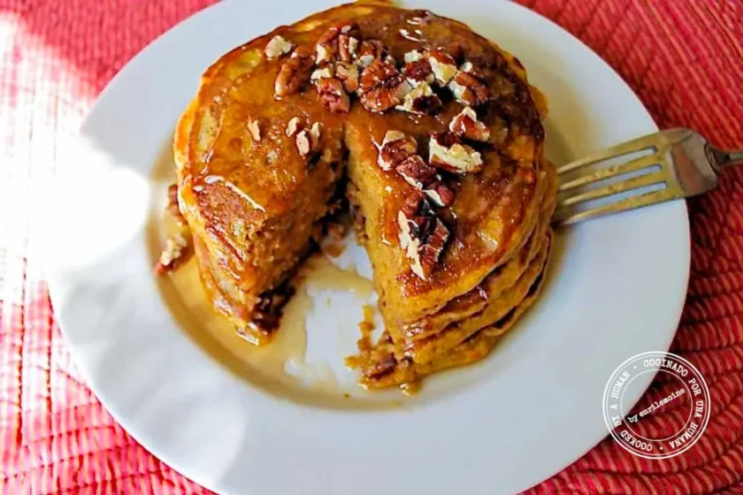 A stack of pancakes topped with pecans and syrup on a white plate, with a fork resting on the side.