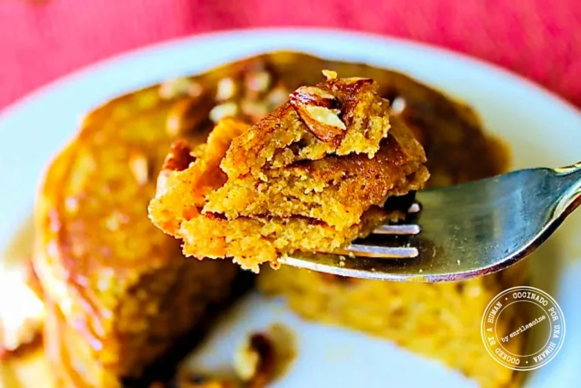 A bite of pumpkin pancake topped with a pecan on a fork, held in front of a stack of pancakes.