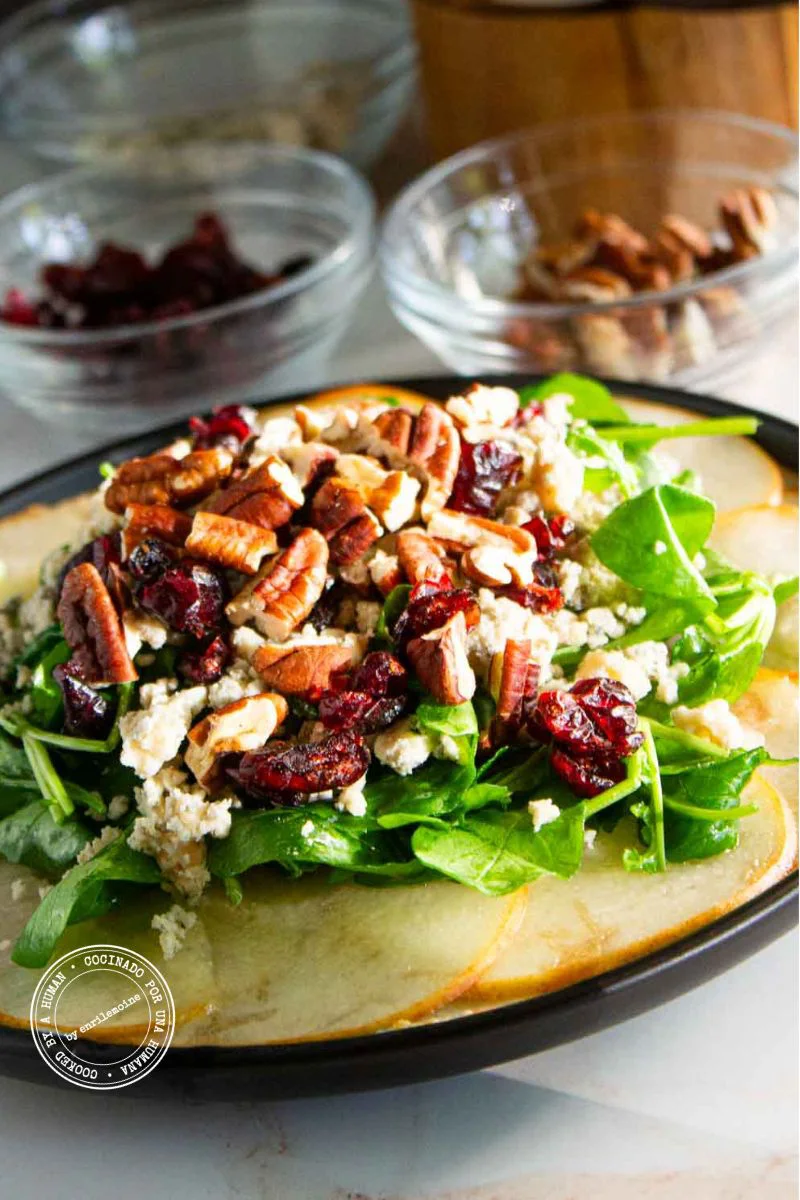 Una ensalada de peras cortadas en rodajas, verduras de hoja verde, arándanos rojos secos, nueces pecanas y queso desmenuzado en un plato.