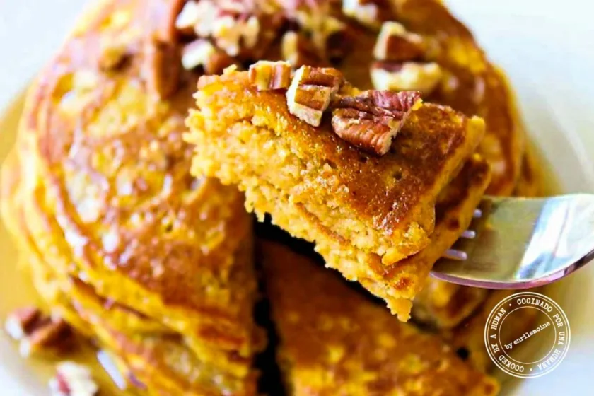 A fork lifting a piece of a pumpkin pancake topped with pecans.