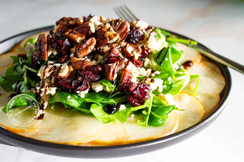 Ensalada con peras cortadas en rodajas, verduras de hoja verde, nueces pecanas, arándanos rojos secos y glaseado de vinagre balsámico, servida en un plato oscuro.