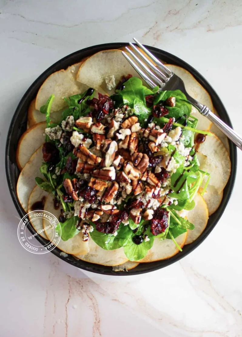 Ensalada con peras cortadas en rodajas, verduras de hoja verde, nueces pecanas, arándanos rojos secos y queso azul, aderezada con un glaseado de vinagre balsámico y decorada con un tenedor plateado.